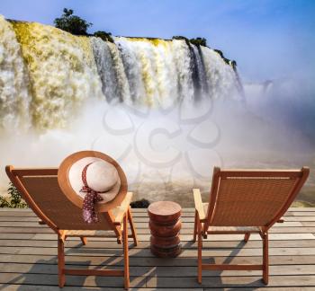  Incredible exotic waterfalls of Iguazu in South America. Two comfortable wooden chaise lounges face waterfalls. On one hangs an straw female hat. Concept of active and eco-tourism