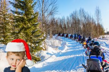 Adorable little boy with blue eyes smiling. He put on a red Santa Claus hat and is waiting for New Year. Train from snowmobiles moves along the ice