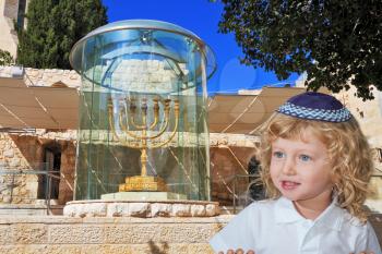 Cute little boy with long blond curls and blue eyes in a knitted skullcap. He stands at the Golden Menorah in Jerusalem. The Jewish holiday of Sukkot