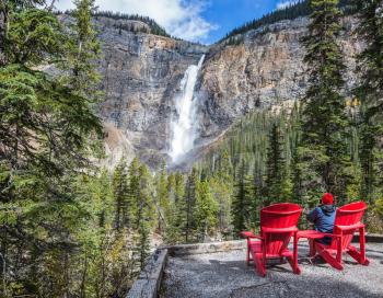 Elderly woman photographs the famous waterfall. Two red deck chairs to relax in front of the waterfall Takakkaw. Autumn day in Yoho National Park in the Rocky Mountains 