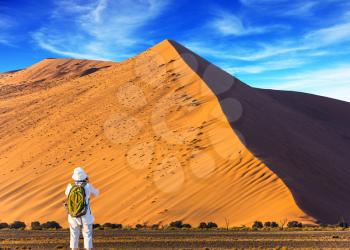 Namib desert. The concept of extreme and exotic tourism. Elderly enthusiastic woman with a green backpack is taking pictures of purple and yellow dunes