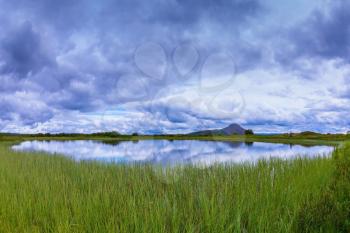  Small lake surrounded by green meadow. Summer Iceland. In the smooth water of cold lake reflects cloudy sky