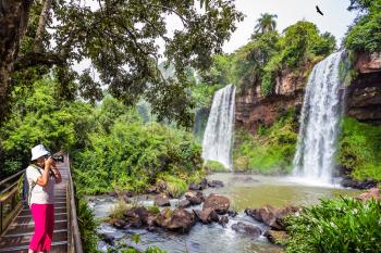 Middle-aged woman in red trousers and panama photographs waterfalls Iguazu Falls. The concept of active and phototourism