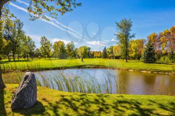 Small pond with a clay bottom is surrounded by reeds. Green grass lawn and trees. Concept of recreational tourism. Shining sunny day in French Canada