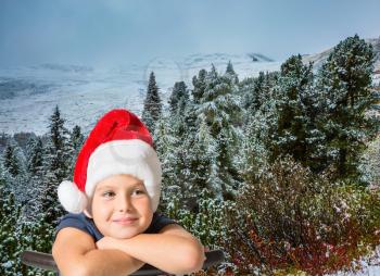  Pine forest in mountains are covered with the first snow. Charming eight-year boy in Santa Claus hat thinking