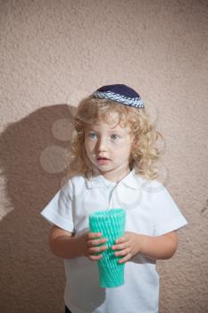 Autumn holiday of Sukkot. Adorable little boy with long blond curls and blue eyes in the Jewish knitted skullcap. He holds the case for the holiday citrus