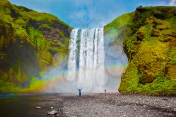 Grand waterfall in Iceland - Skogafoss. Picturesque huge rainbow appears in the water mist. Middle-aged woman - tourist shocked beauty waterfall
