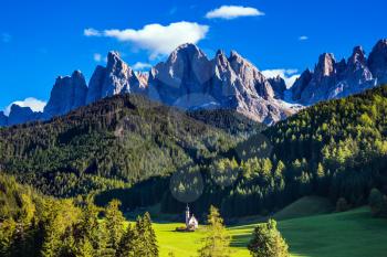 Dolomites, Tirol. Sunny warm autumn day. The famous symbol of the valley Val di Funes - church of Santa Maddalena. Rocky peaks and forested mountains surrounded by green Alpine meadows
