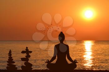 Silhouette of yogi in lotus position and a piles of stones at sea shore at sunrise