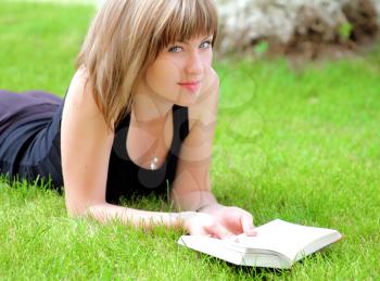 Beautiful young smiling woman laying on grass reading book, against green of summer park.