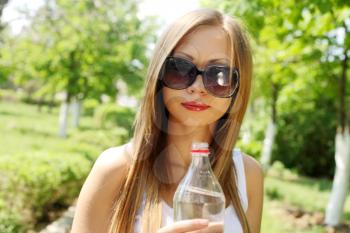 Portrait of young beautiful blond-haired woman wearing white t-shirt drinking water at summer green park.