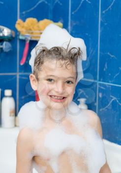 Smiling little boy front view. Kid covered with soap bubbles against a blue wall in bathroom