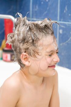 Smiling little boy front view. Kid covered with soap bubbles against a blue wall in bathroom. Closed eyes