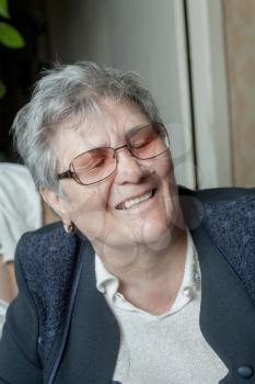 Closeup portrait of senior woman with glasses, looking at camera, smiling. Indoors. Closed eyes.