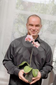 Handsome caucasian 40s man smiling portrait on grey background with black shirt holding orchid in pot. A business man having a blooming pink orchid