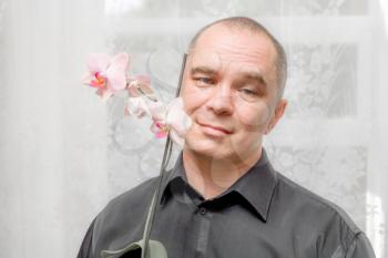 Handsome caucasian 40s man smiling portrait on grey background with black shirt holding orchid in pot. Man with domestic flowers looking at camera, blooming orchid