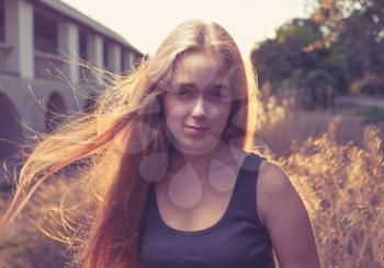 Long blond hair fly by the wind. Young blonde women at sunset backlit. Blank expression on her pretty young face. Looking at camera very confident.