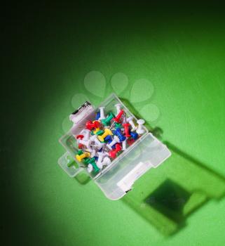 image of mixed colors office push-pins in the plastic pack, angle view, on the green background, natural vignette made by flash light