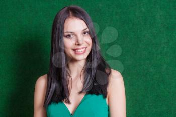 Cute lady looking at camera, head and shoulders shot. Brunette weared green dress smiling against green wall