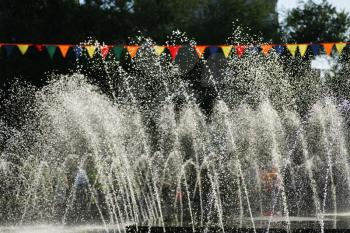 Rows of fountain water jets spraying water into the air in a public park on a sunny summer day and decoratoon of red flags over