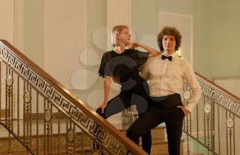 Fashionable couple, woman in party blouse and man in shirt with bow tie posing on stairs inside fancy mancion toned image. Woman leanin on mans shoulder and looking at him