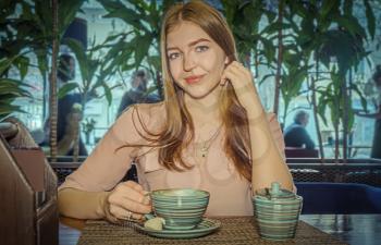 Smiling blonde touching her hair by one hand and cup of coffe by another. She is relaxing in cafeteria with mug of strong coffee in coffee-break