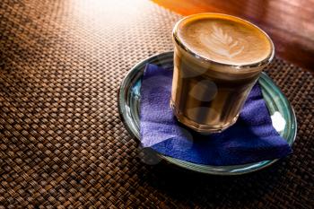 Flat white coffee froth art on top is served on saucer over the table with bamboo mat backlit, copyspace