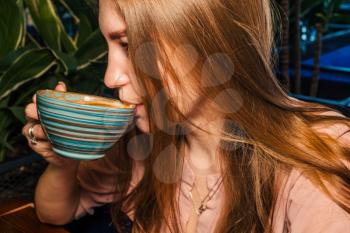 Side view of beautiful lady lond blond hair drinking from cup of coffee in hands. Cropped Image Of lass Having Coffee Drink