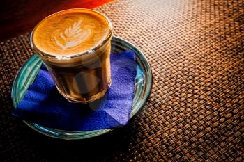 Flat white coffee is served on saucer over the table with bamboo mat backlit, copyspace