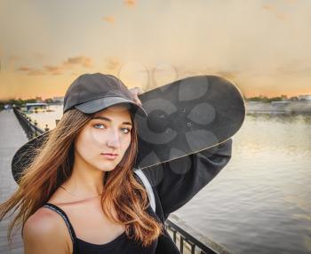 A young woman wears a T-shirt with straps and a black hat, she holds a skateboard on her shoulder and looks at the camera. Loneliness in town. The river and the sunset sky in the background.
