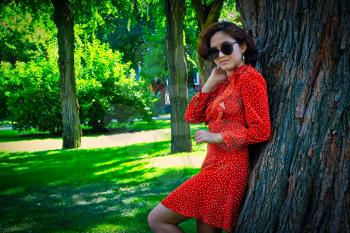 Happy girl in sunglasses dressed in red summer dress posing leaning park tree in summertime, copyspace