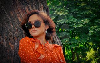 Nice Asian girl poses in retro sunglasses leaning against the trunk of a huge tree in the forest. She is dressed in a vintage dress of red colour and on the right part of image there is plenty of room for text.
