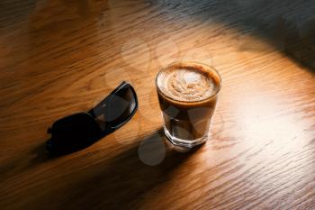 Flat-white coffee in small glass served on table with sunglasses lying near on wooden tabletop.