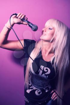 Blonde model lady karaoke singer with a microphone over pink violet background, side view.
