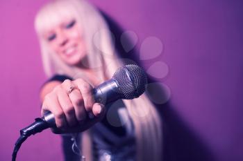 Blonde lady karaoke singer with a microphone over pink violet background outstretched hand towards the camera, blurred background, focus on microphone, copyspace mock-up banner