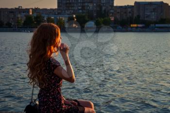 Relaxed redheaded lady sitting on the river side in setting sun light, side view.