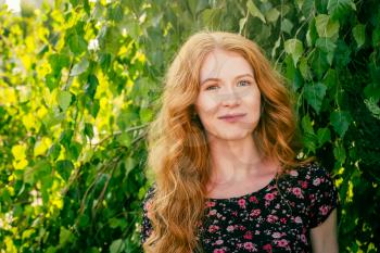 Close-up portrait of lovely lady with long red ginger hair and freckles smiling looking at camera in park in summer. People in nature and happiness concept.