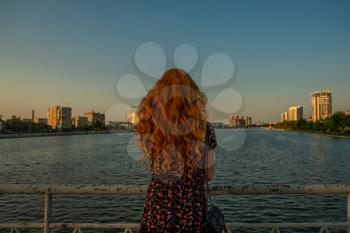 Sad redhead woman silhouette standing alone on a bridge in autumnal sunset looking at city urban view.