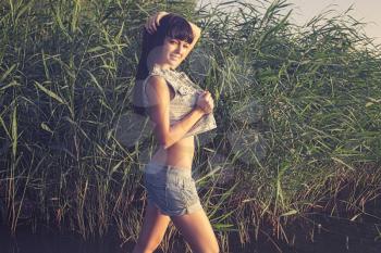 Brunette slim lady posing in denim shorts in front of reeds in sunlight alone.