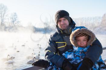 Man with son at winter nonfreezing lake with white whooping swans. The place of wintering of swans, Altay, Siberia, Russia.