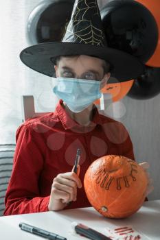 Teen boy in costume preparing for the Halloween celebration drawing a pumpkin. Halloween carnival with new reality with pandemic concept. Children wearing face masks to protect against COVID-19.
