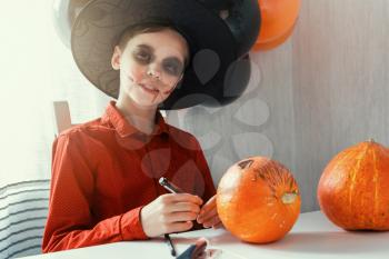 Happy teen boy in costume preparing for the Halloween celebration drawing a pumpkin. Halloween carnival or masquerade concept