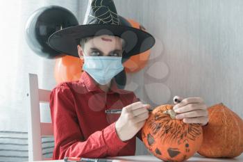 Teen boy in costume preparing for the Halloween celebration drawing a pumpkin. Halloween carnival with new reality with pandemic concept. Children wearing face masks to protect against COVID-19.