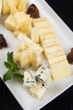 Cheese plate on a dark background table. Many kinds of cheese with sauce and greens on a white plate, closeup shot