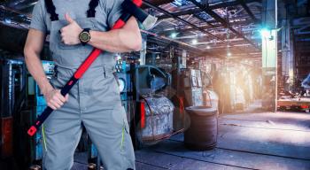 Closeup of worker man with ax dressed in overalls on Industrial zone background, manufacturing facility. Concept of Safe labor in the enterprise.