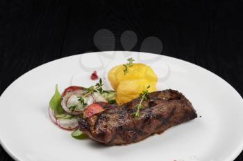 Grilled beef skirt steak meat with potato and vegetables on white plate on wooden black background