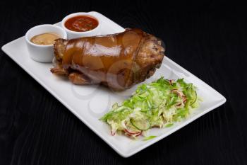 Tasty pork knuckle with sauces and vegetables on a black wooden background