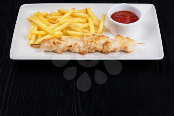 Grilled chicken shashlik meat with fried potatoes on white plate on black wooden background