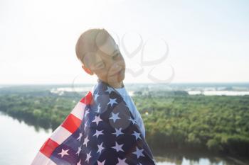 Blonde boy waving national USA flag outdoors over blue sky at the river bank. Beauty summer sunny day. American flag, patriotism, independence day 4th july concept