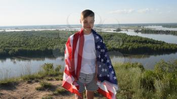 Blonde boy jumping and smiling with waving national USA flag outdoors over blue sky at the river bank. Beauty summer sunny day. American flag, patriotism, independence day 4th july concept
